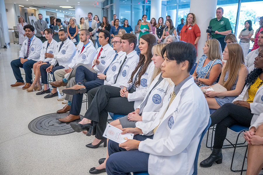 A group of medical students sit on the front row of a large group of people during a ceremony recognizing them for an achievement.