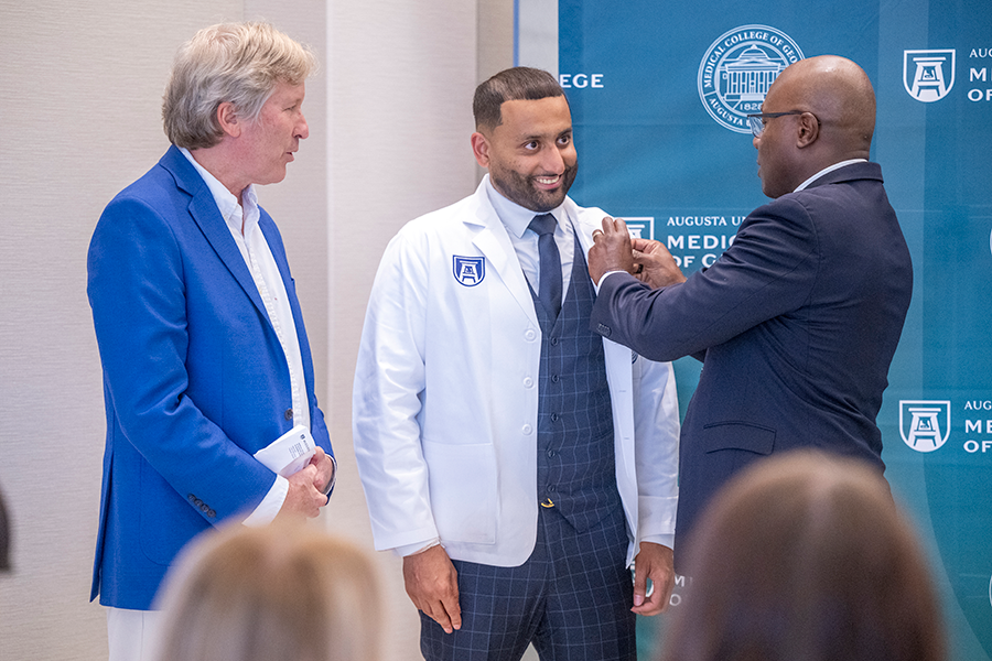 A male medical student has a pin placed on his lab coat by two other men during a ceremony.