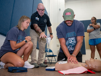 Three college athletics coaches learn how to administer CPR using a dummy while a CPR trainer gives instructions.