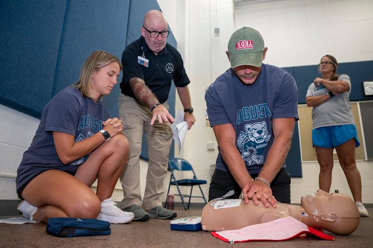 Three college athletics coaches learn how to administer CPR using a dummy while a CPR trainer gives instructions.