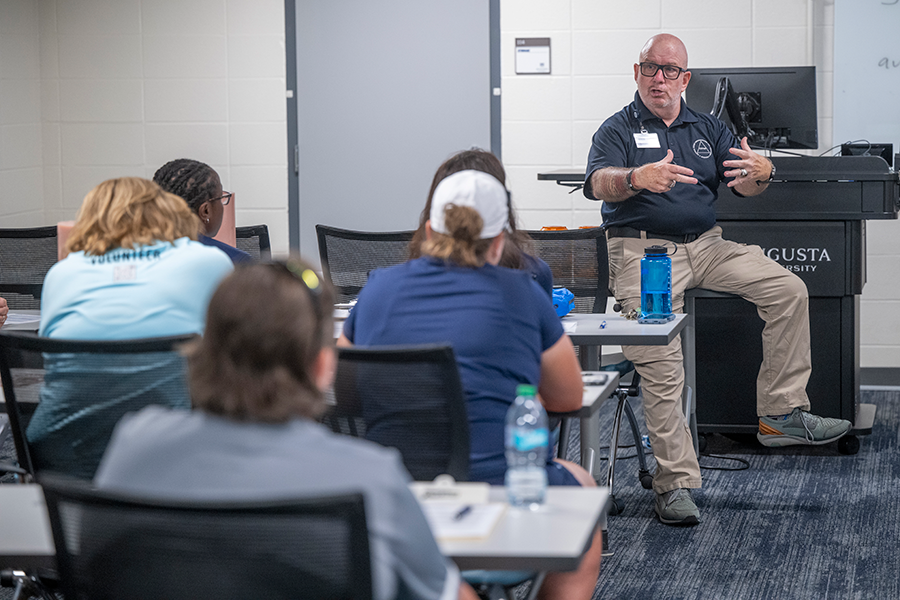 A man talks to a room full of college athletics coaches about the importance of being trained in CPR and Stop the Bleed.