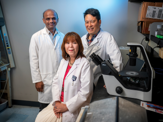 Two male and one female doctor in lab coats sit in front of a microscope in a laboratory and smile at the camera.