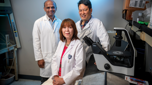 Two male and one female doctor in lab coats sit in front of a microscope in a laboratory and smile at the camera.