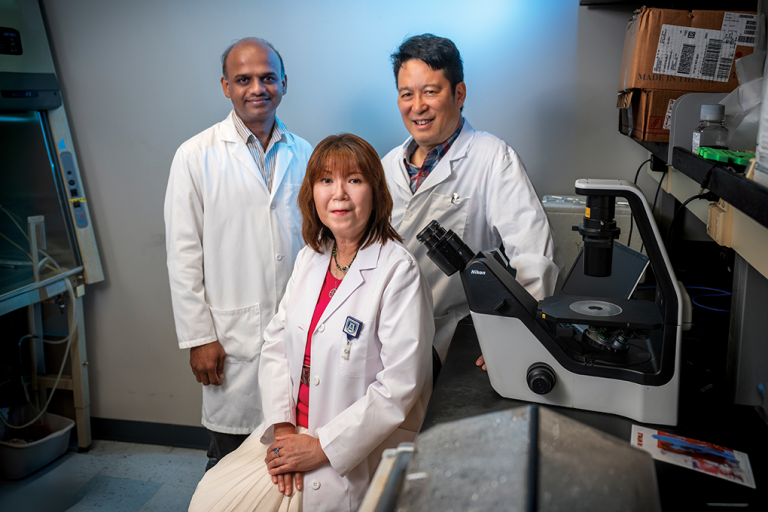 Two male and one female doctor in lab coats sit in front of a microscope in a laboratory and smile at the camera.