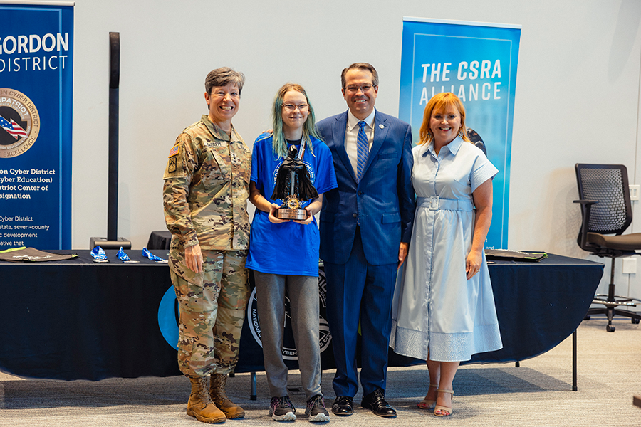 Two women and a man stand with a female high school student holding an award. One woman is a soldier in fatigues.