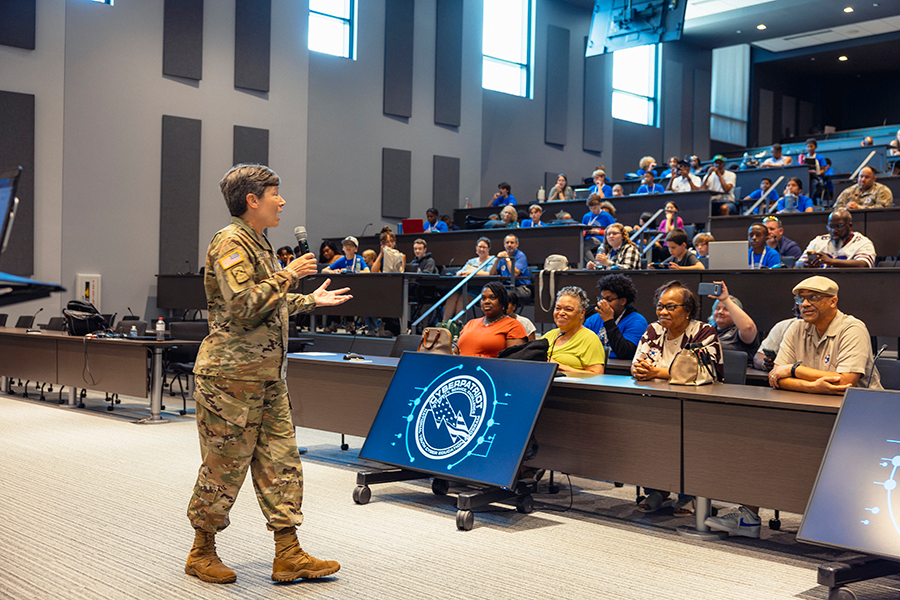 A female soldier in fatigues speaks to a large crowd in a large auditorium.