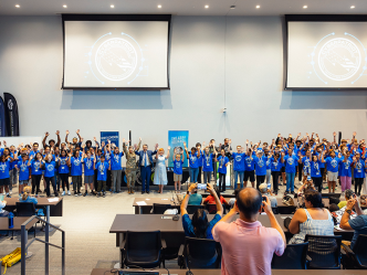 A large group of close to 100 high school students cheer to celebrate completing a summer camp. They are at the front of a large auditorium while their parents and others cheer and take pictures.