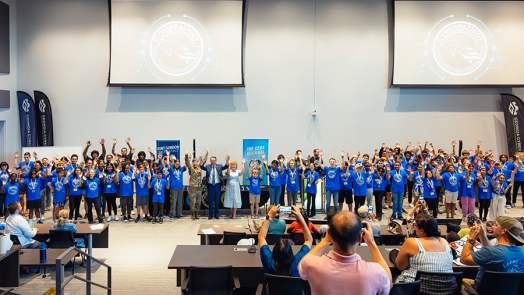 A large group of close to 100 high school students cheer to celebrate completing a summer camp. They are at the front of a large auditorium while their parents and others cheer and take pictures.