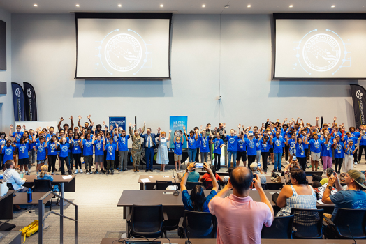 A large group of close to 100 high school students cheer to celebrate completing a summer camp. They are at the front of a large auditorium while their parents and others cheer and take pictures.
