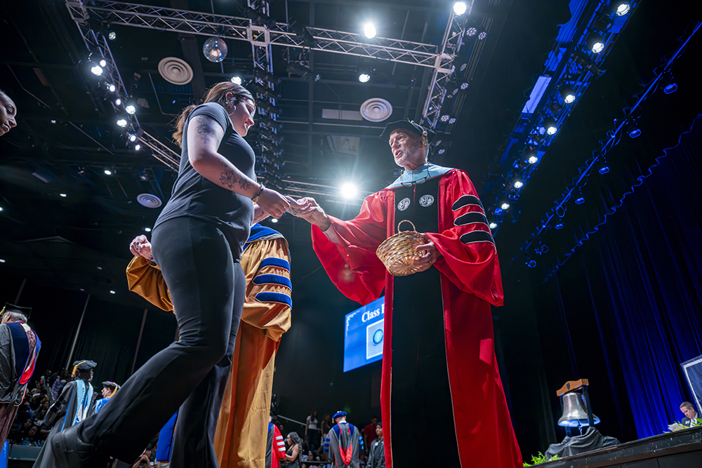 man in a cap and gown hands a female student something from a wicker basket.