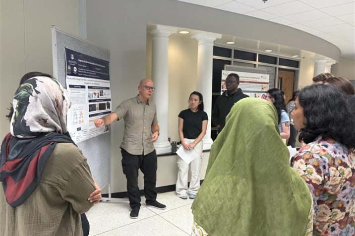 A man in a grey shirt and glasses, Dr. Paul Langridge, presents a research poster to a crowd of PhD students.