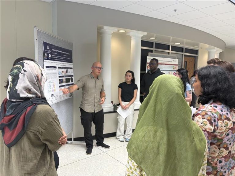 A man in a grey shirt and glasses, Dr. Paul Langridge, presents a research poster to a crowd of PhD students.
