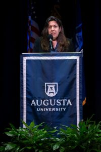A woman stands at a podium and addresses a large group of college students.
