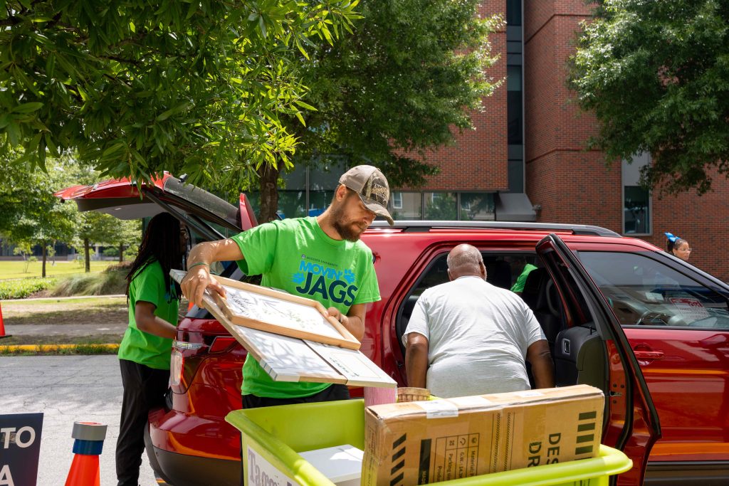 Several people are gathered around a vehicle while loading personal belongings into a large box.
