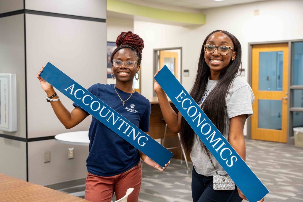 Two college-aged female students inside a classroom are holding up two signs.