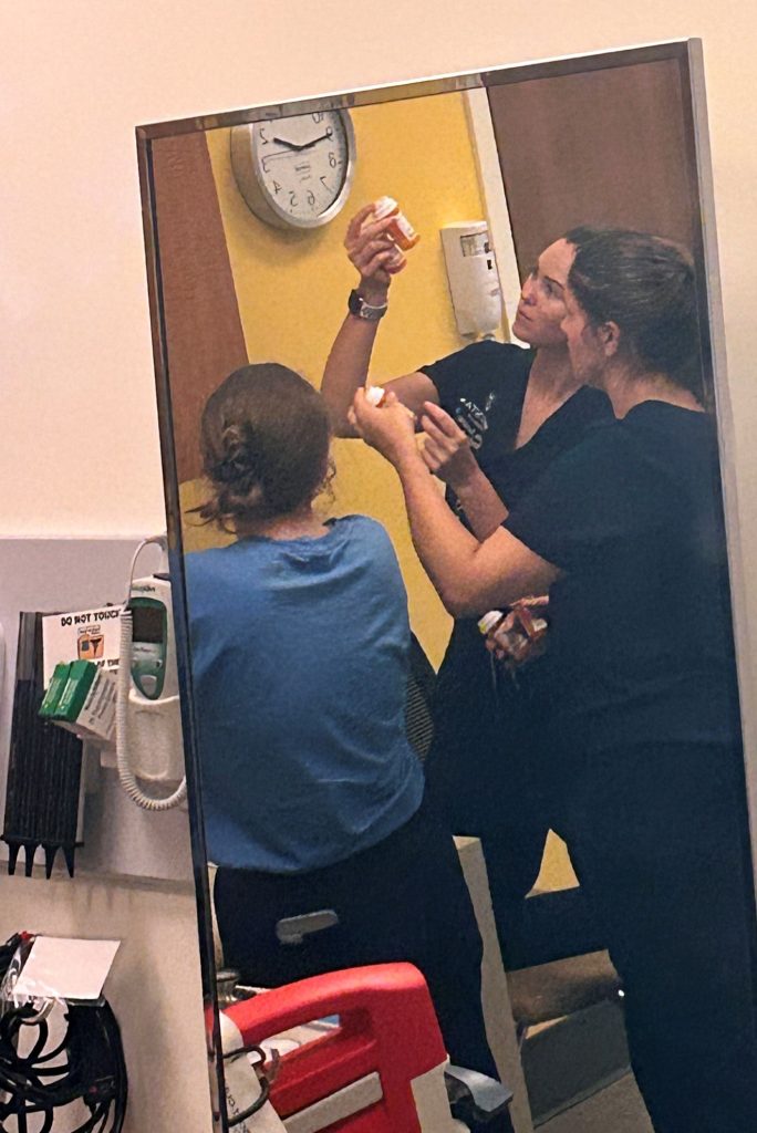 Three female students gather together to look at a pill bottle, as seen via a mirror in the room