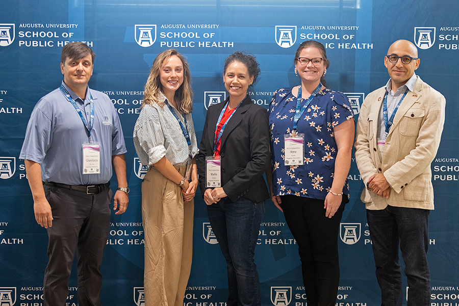 Three women and two men stand in front of a large backdrop that features the logo for Augusta University's School of Public Health.