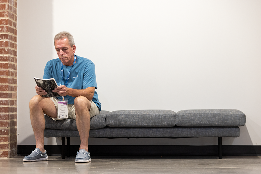 A man sits on a bench inside a building. He is holding a notebook and going over his notes for a presentation.