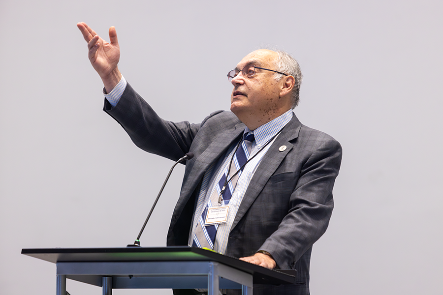 A man stands at a podium with a microphone and raises his right arm while addressing a large group of people inside an auditorium.