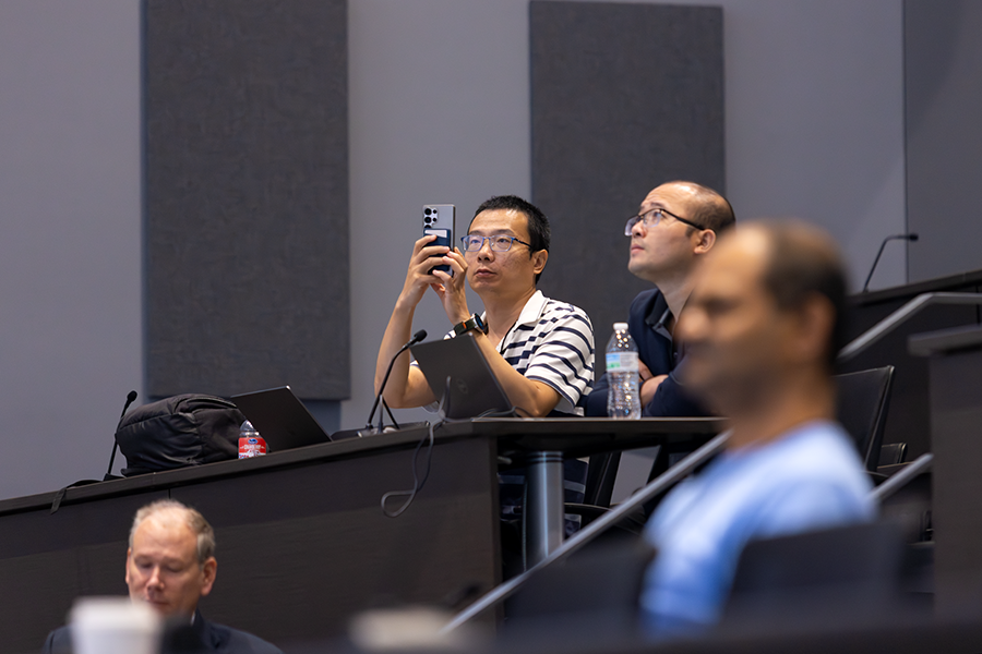 A man sitting in a crowded auditorium takes a picture of a screen with his cellphone.