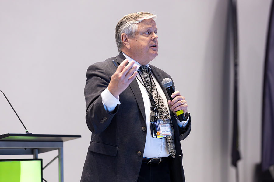 A man holds a microphone while speaking to a large group of people gathered in an auditorium.