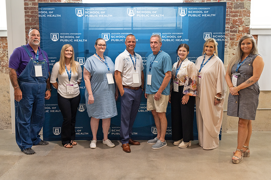 A group of eight people, five women and three men, stand in front of a banner prior to giving a series of presentations. The banner has the logo for Augusta University's School of Public Health on it in several places.
