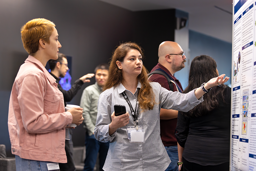 A college woman speaks with another woman and talks about her scientific poster. She is pointing to one section of the poster.