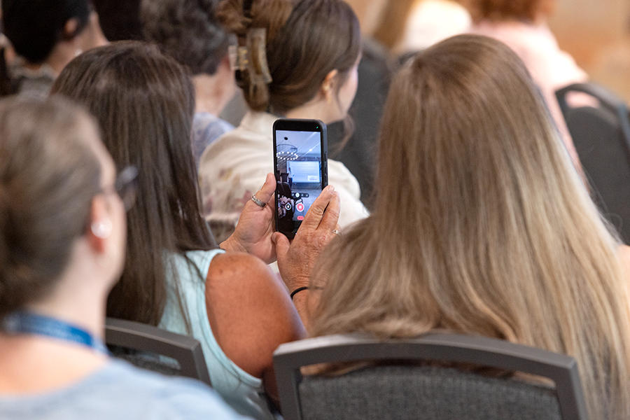 A woman holds up her phone to take a video of her husband giving a presentation in front of a room full of people.