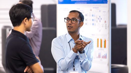 Two college men talk during a poster session for a symposium. One man is using his hands to indicate what he is describing.