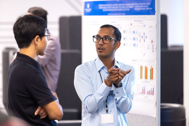 Two college men talk during a poster session for a symposium. One man is using his hands to indicate what he is describing.