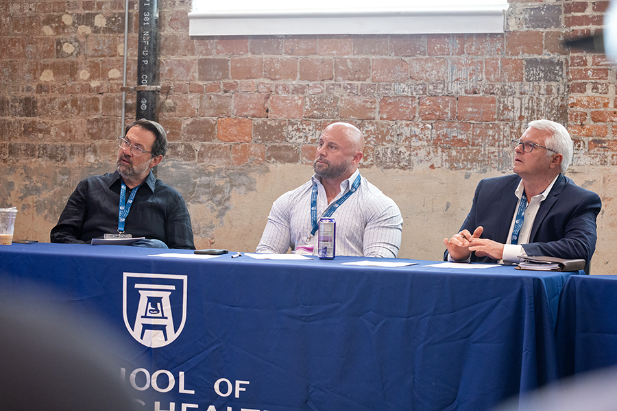 Three men sit at a table and listen to a series of presentations.