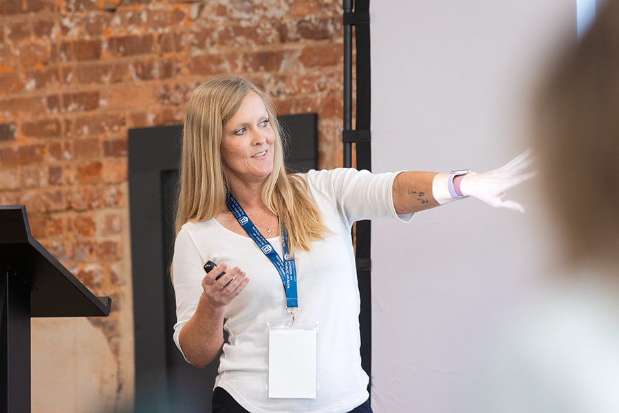 A woman gives a presentation to a large group of people gathered in a conference room. She is pointing to a slide on a projector screen.