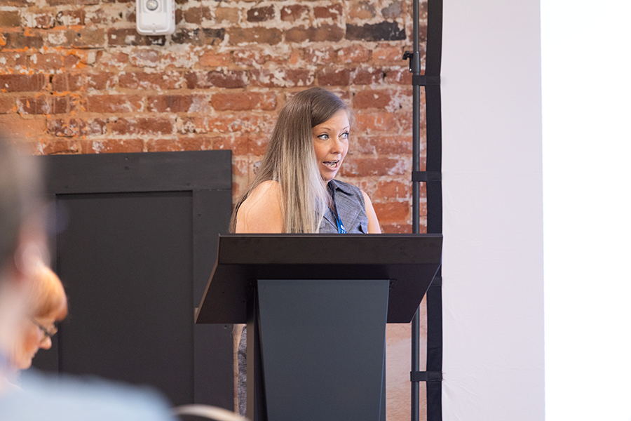 A woman stands at a podium and gives a presentation to a large group of people gathered in a conference room.