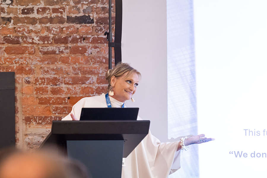 A woman stands at a podium and gives a presentation to a large group of people gathered in a conference room.