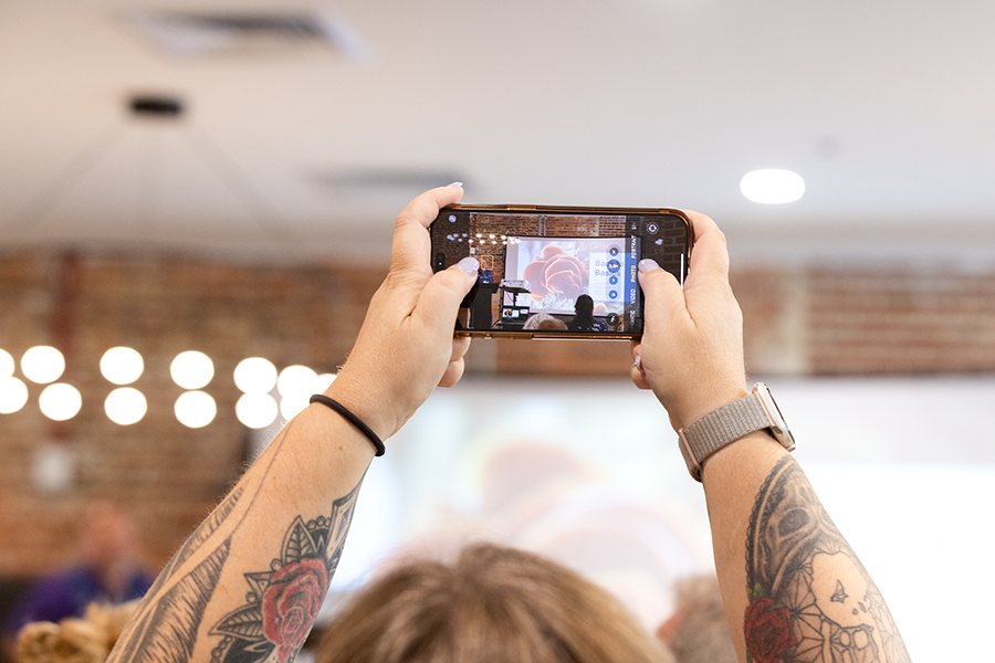 A woman holds her phone over her head to take photos of her husband presenting in front of a large crowd.
