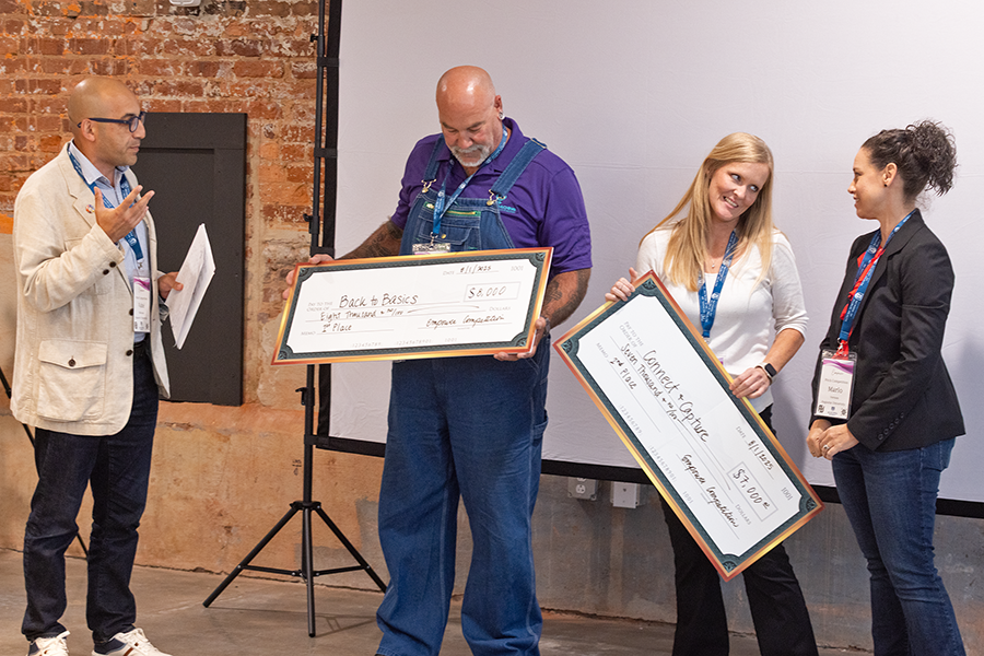 A man and a woman present oversized checks to another man and woman. The man who won has a check that symbolizes an $8,000 investment into his company. The woman who received her check is holding one for $7,000.