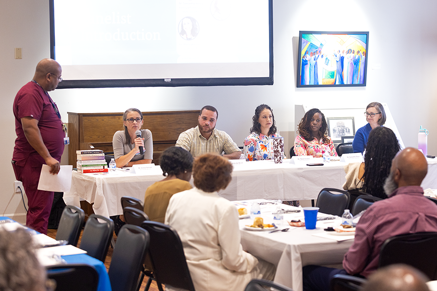 A panel of five researchers, including four women and one man, sit at a table and speak to a large group of people.