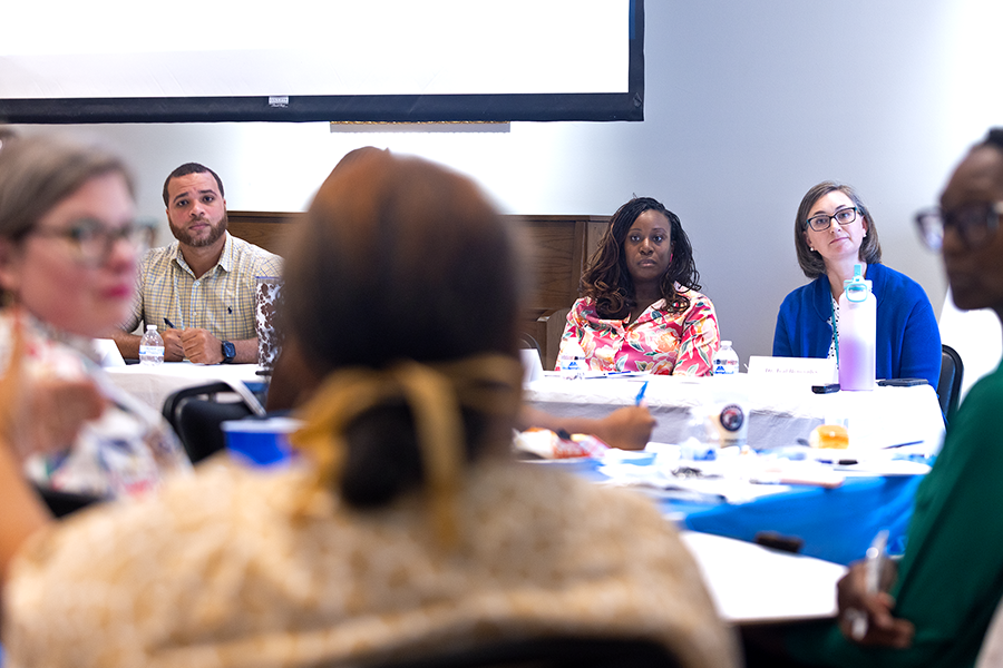 Three people, two women and one man, sit at a table at the front of a room and listen as a woman in a large crowd speaks to them.