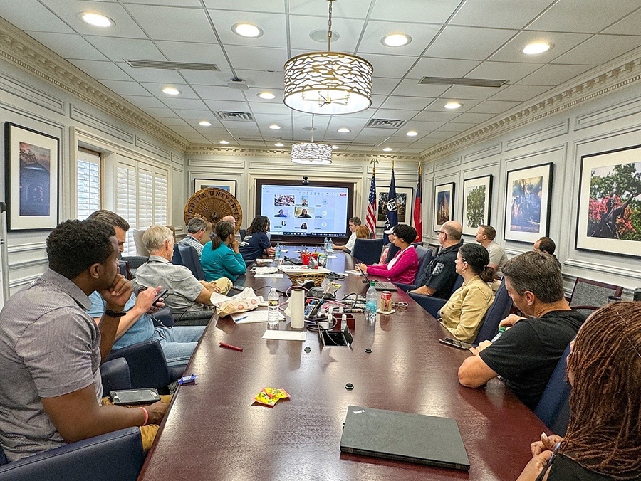 A large group of people sit around a conference table. There is a large TV screen at one end of the room with more pepple gathered online.