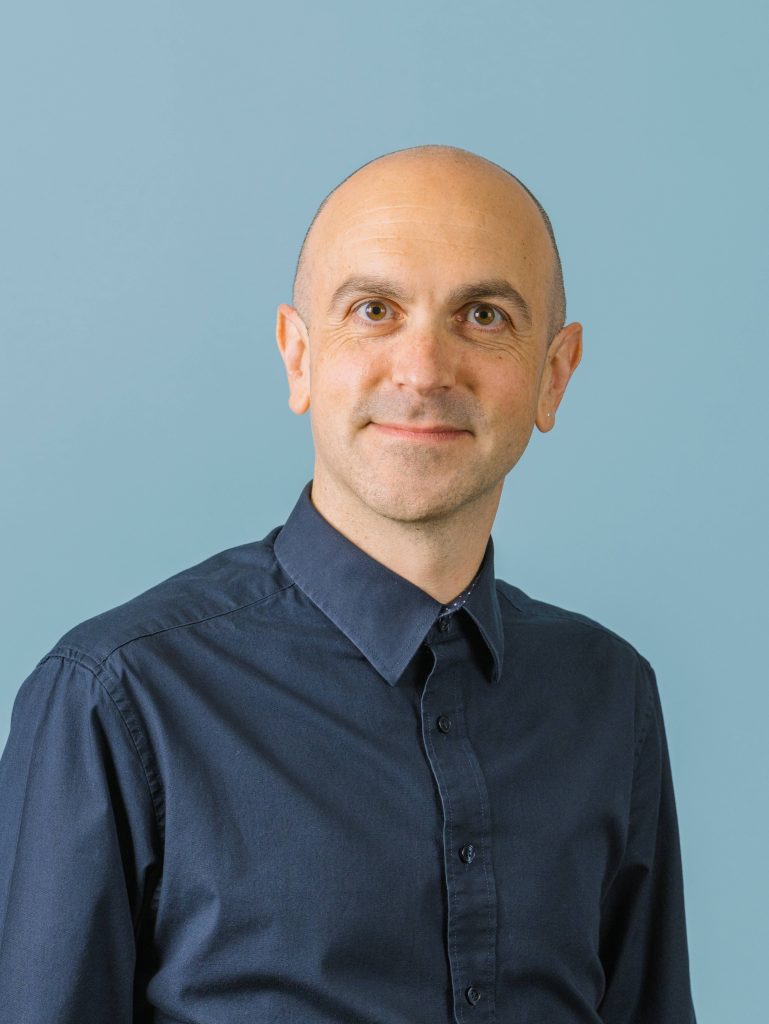 Headshot of a man in a button up dress shirt. He is smiling at the camera and is on a solid, lighter background.