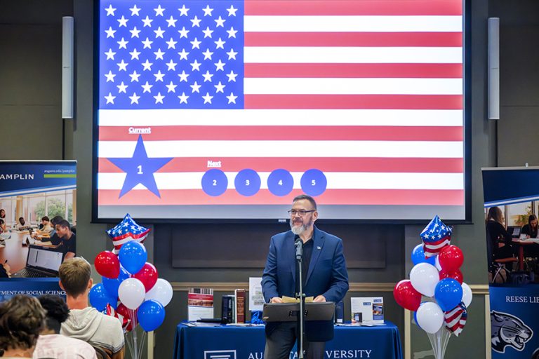 A man stands in front of a large crowd with an American flag behind him.