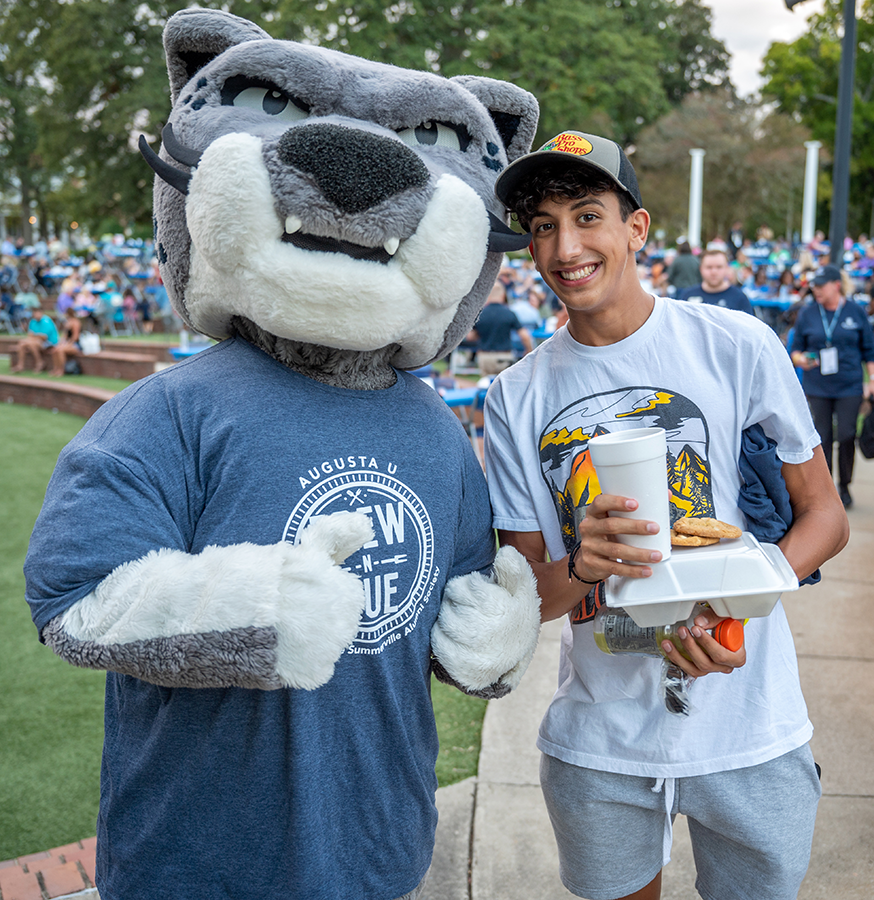 A college mascot modeled after a jaguar stands with a male college student holding a to-go box of food and a drink.