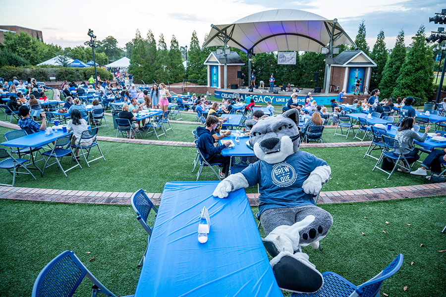 A college mascot modeled after a jaguar sits at a picnic table as a large group of people eat at similar tables spread around an outdoor amphitheater. There is a band setting up on a stage behind the mascot.
