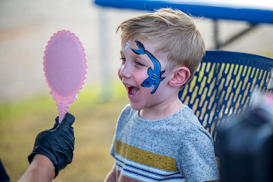 A young boy smiles as he looks at himself in a small mirror. He has a shark painted on his face.