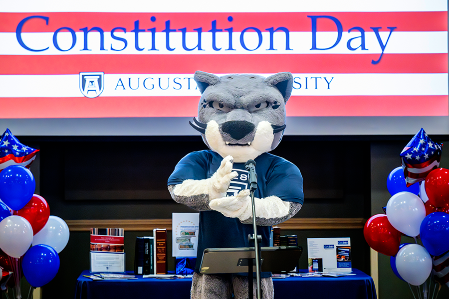 A college mascot modeled after a large cat stands in front of a large crowd at a microphone and reads part of the U.S. Constitution.