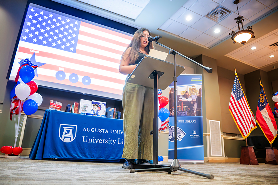 A female college student stands in front of a large crowd at a microphone and reads part of the U.S. Constitution.