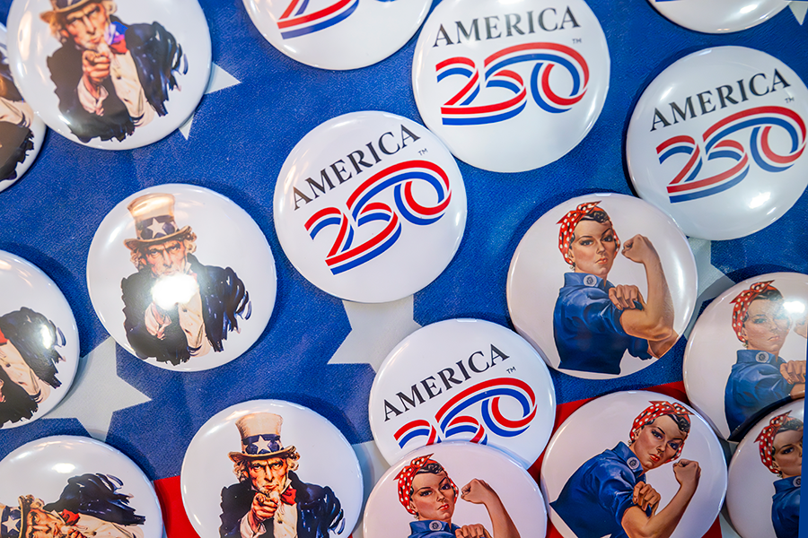 Buttons on a table. Some have an old man wearing a top hat, some have a woman flexing her muscle, and others have "America 250" printed on them.