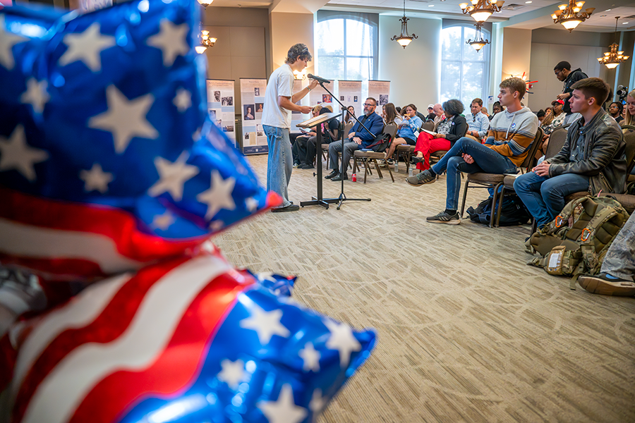 A male college student stands in front of a large crowd at a microphone and reads part of the U.S. Constitution.