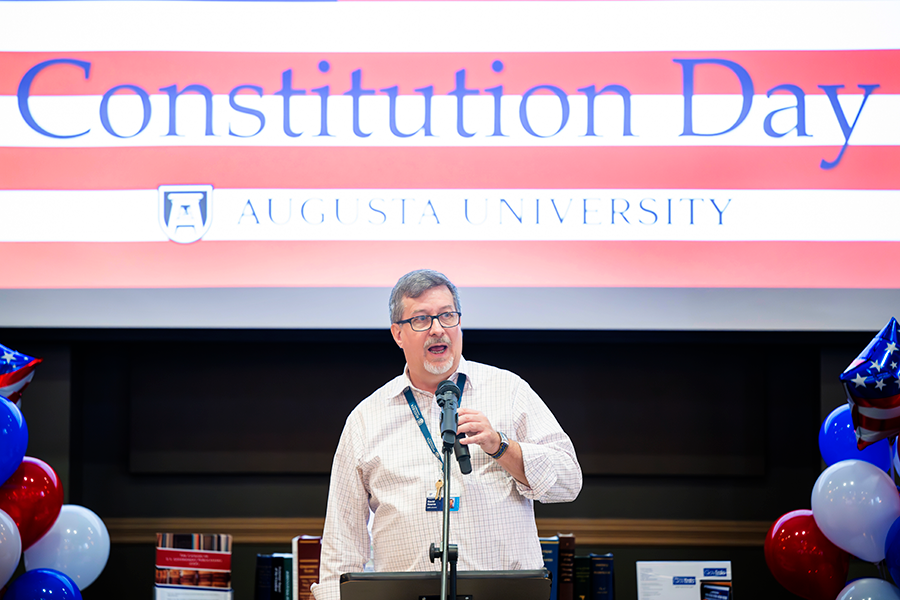 A man stands in front of a large crowd at a microphone and reads part of the U.S. Constitution. Behind him are the words "Constitution Day Augusta University."