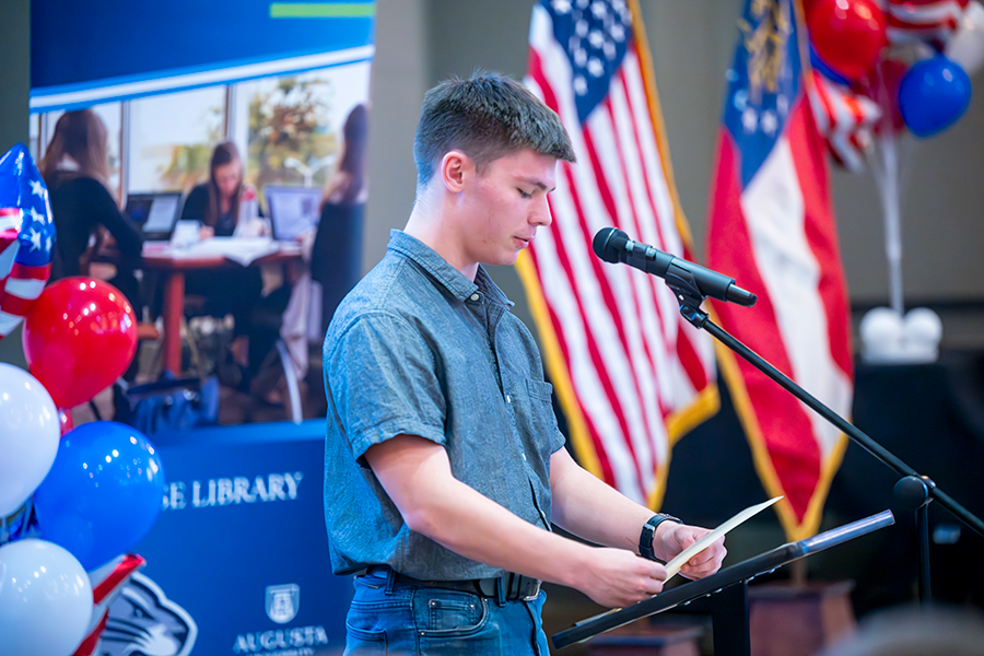 A male college student stands in front of a large crowd at a microphone and reads part of the U.S. Constitution.